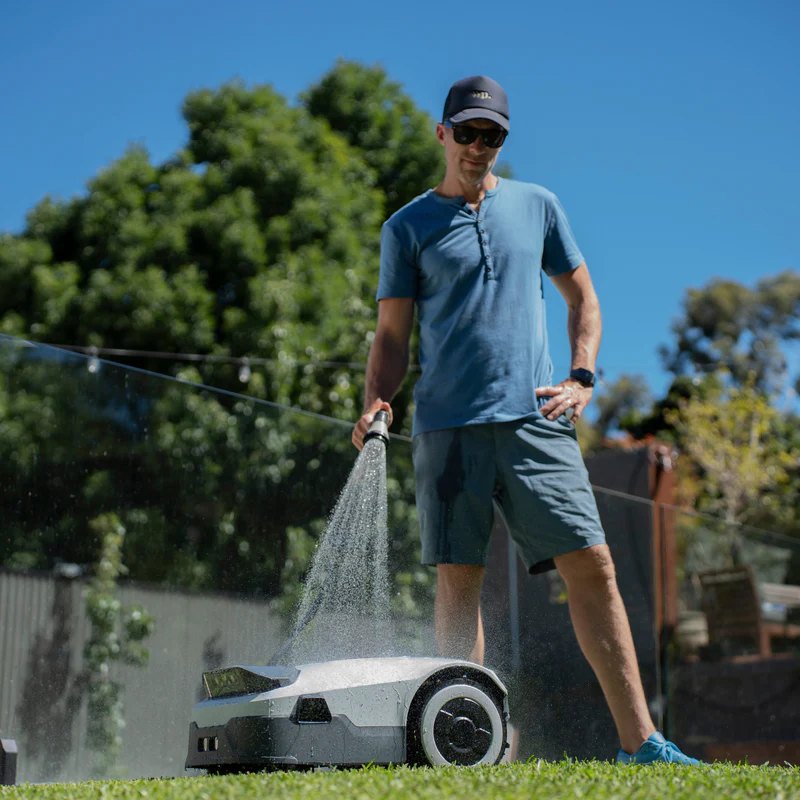 Man cleaning a modern robotic lawn mower with a water spray on a green lawn – maintenance of smart gardening technology for lawn care.