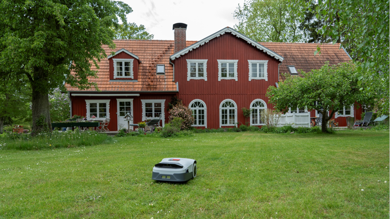 A robotic lawn mower sits stationary on an expansive green lawn in front of a traditional red Scandinavian-style house surrounded by mature trees.